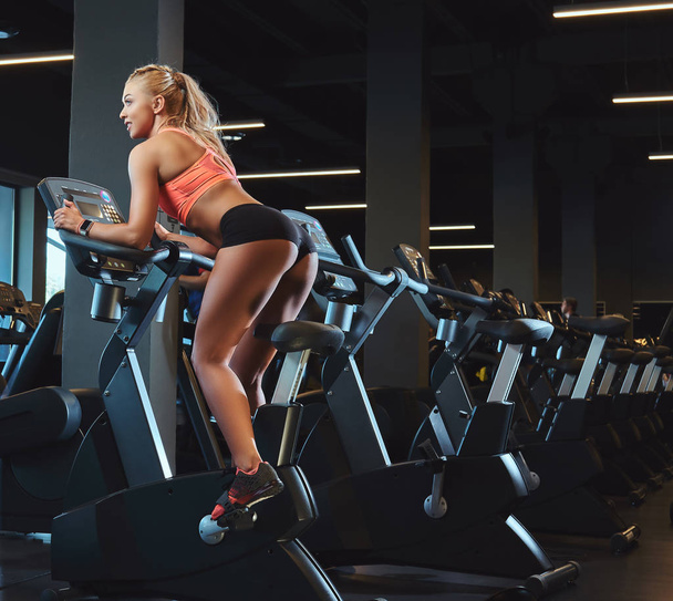 Молодая красивая блондинка в спортивной форме тренируется на велотренажере в современном фитнес-центре
. - Фото, изображение