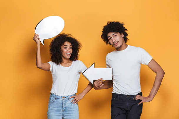 Portrait of an excited young afro american couple holding empty speech bubble and pointing with arrow isolated over yellow background - Foto, imagen