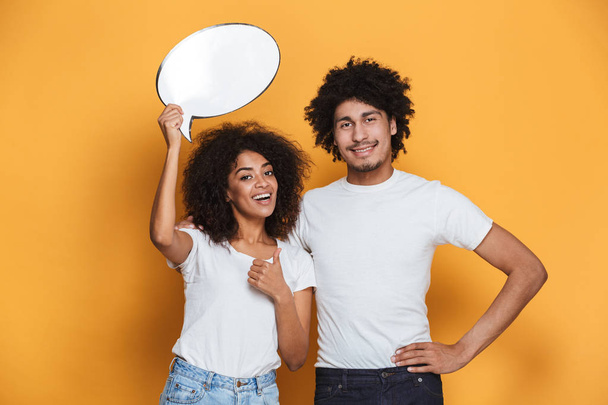 Portrait of a happy young afro american couple holding empty speech bubble isolated over yellow background - 写真・画像