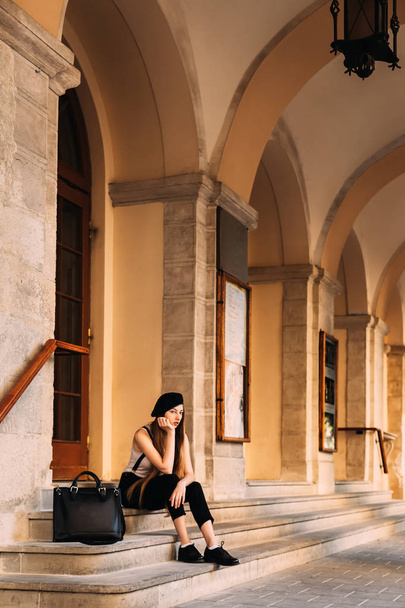 menina pensativa com cabelos longos e roupas elegantes sentado em uma escada de pedra e uma bolsa preta perto dela
 - Foto, Imagem