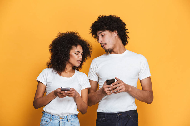 Portrait of a suspicious afro american couple with mobile phone isolated over yellow background - Zdjęcie, obraz