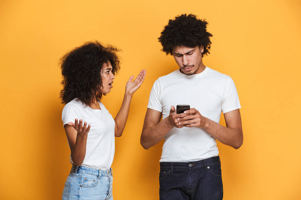 Retrato de una joven pareja afro-americana frustrada discutiendo por teléfono móvil aislado sobre fondo amarillo
 - Foto, imagen