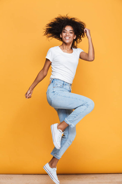 Full length portrait of a joyful young african woman celebrating success while jumping isolated over yellow background - 写真・画像