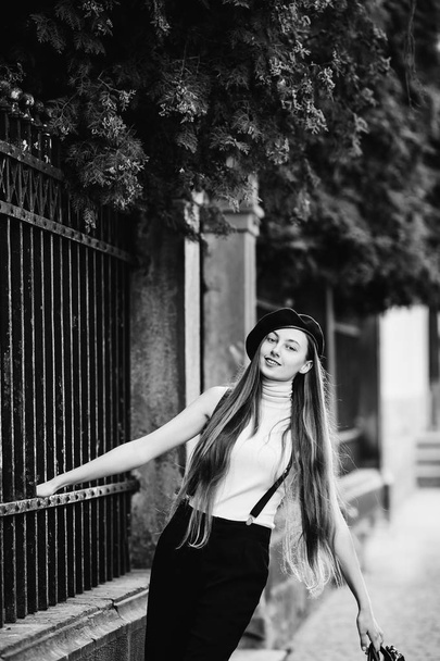 A stylishly dressed girl stands on the sidewalk while clinging to the fence and smile - Fotografie, Obrázek