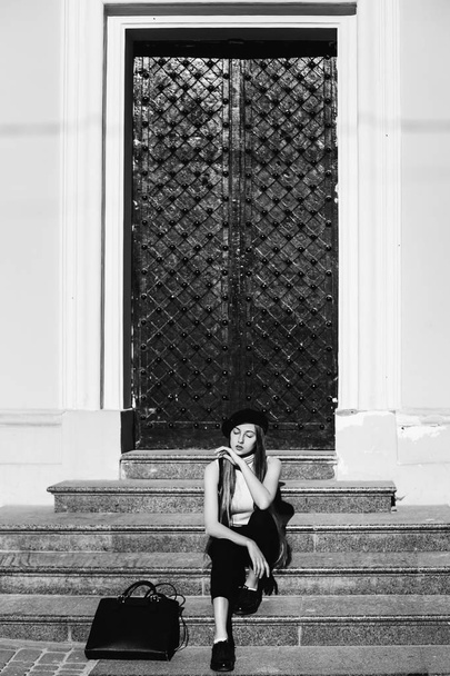 mooi meisje zittend op de trappen van een oud huis met metalen deuren op zwart-wit fotografie - Foto, afbeelding