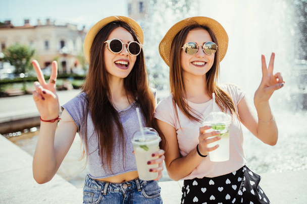 zwei glückliche schöne Mädchen trinken lustige Cocktails, während sie im sonnigen Sommertag mit Friedensgeste in der Nähe des Brunnens spazieren gehen. - Foto, Bild