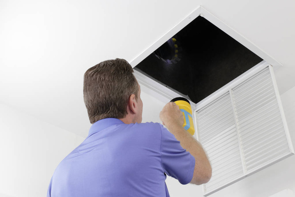 Man Inspecting an Air Duct with a Flashlight. Older male with a yellow flashlight examining HVAC ducts in a large square vent. Male technician looking over the air ducts inside a home air intake vent. - Photo, Image