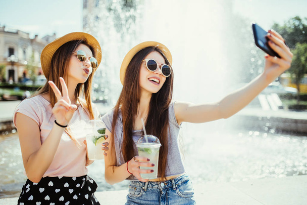 Дві щасливі жінки в сонцезахисних окулярах і капелюхах гарно проводять час разом у місті, беручи селфі біля фонтану на вівтарі
 - Фото, зображення