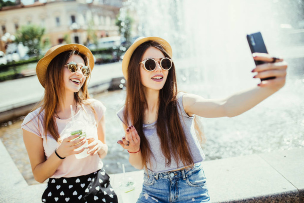 Дві жінки найкращі друзі відновлюються разом проти фонтану, радісно дивляться на сучасний смартфон, спілкуються з друзями, підключені до бездротового Інтернету та п'ють свіжі коктейлі
 - Фото, зображення