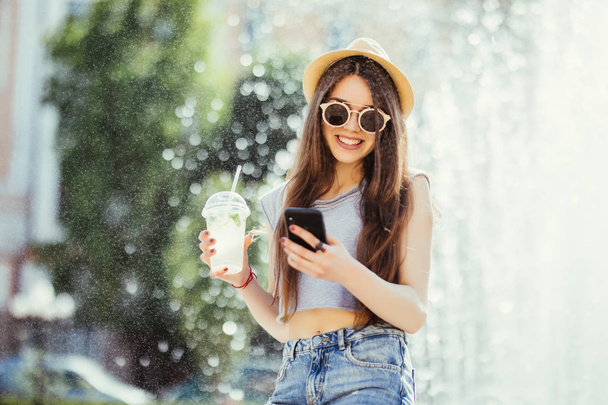 Young woman in summer outfit drink cocktails outdoors typing on phone near fountain - Zdjęcie, obraz