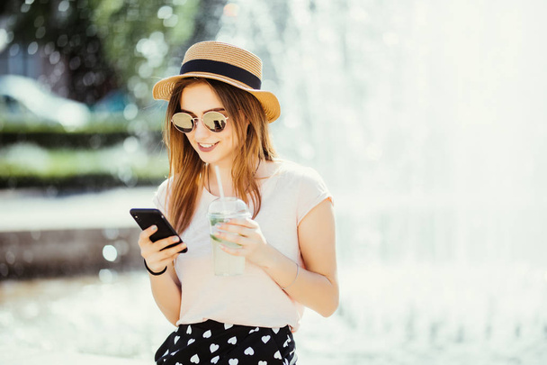 Jovem mulher de beleza de verão com telefone conversando ou surfar na internet bebe mojito coquetel de frutas contra fonte na rua
 - Foto, Imagem