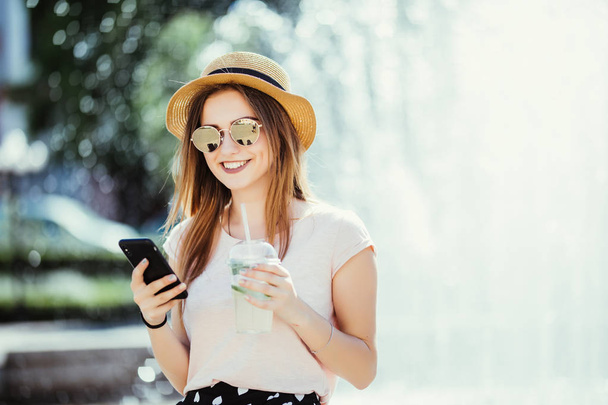 Jonge mooie brunette vrouw met een koud drankje zitten in de straat te typen op de telefoon - Foto, afbeelding