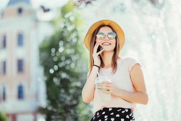 Όμορφη νεαρή γυναίκα σε γυαλιά ηλίου και το καπέλο σε καφέ πίνοντας mojito δροσιστικά κοκτέιλ ενώ μιλάτε στο τηλέφωνο ηλιόλουστη ημέρα - Φωτογραφία, εικόνα