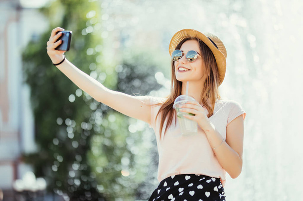 Молодая девушка делает селфи из рук с телефоном на летней улице города. Концепция городской жизни
. - Фото, изображение