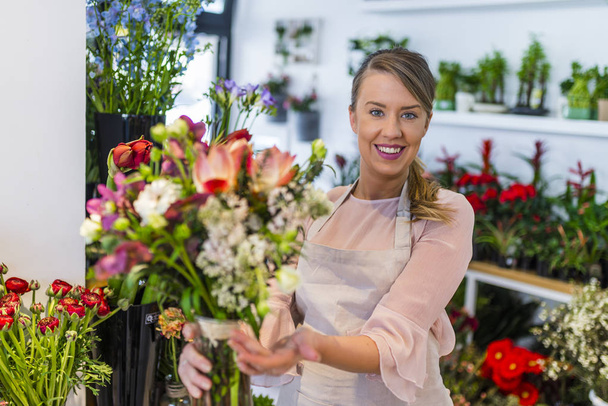 Portrait of a florist offering flowers at the counter in the florist shop. Young woman giving bouquet of flowers. Portrait of a happy florist offering and sale flowers at the counter in the florist shop. Florist giving beautiful flower bouquet to cli - Zdjęcie, obraz