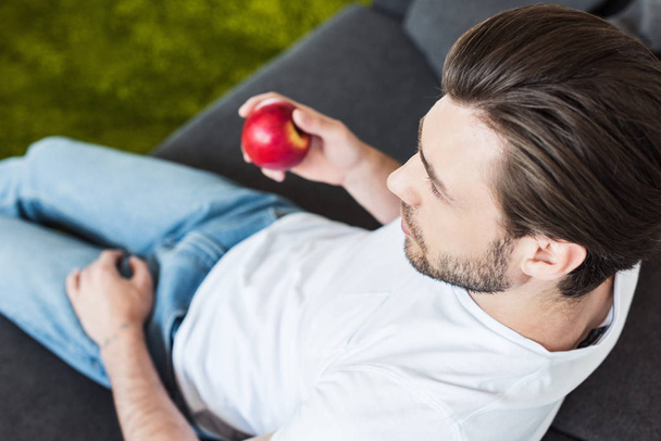 high angle view of young man sitting on sofa with apple in hand at home - Фото, изображение
