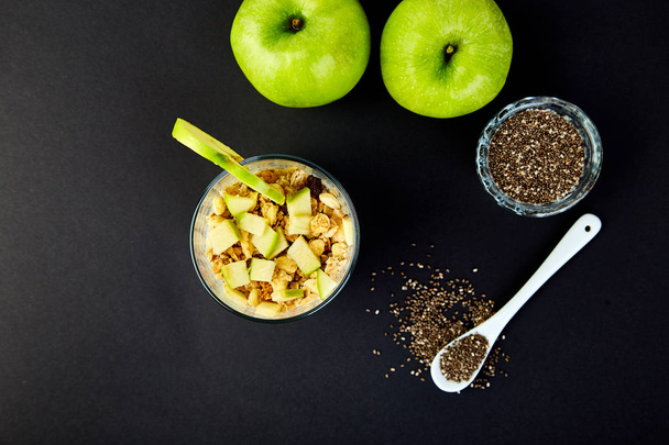 Healthy chia pudding with apples and granola in glass on black background. Vegan creamy breakfast.Detox and healthy superfoods breakfast concept. Overhead, top view, flat lay. - Photo, Image