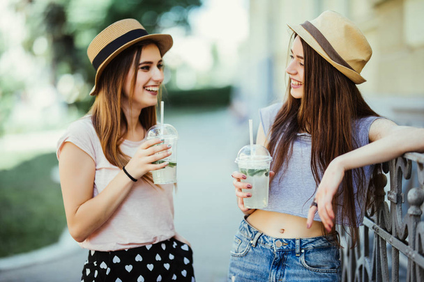 Две молодые женщины разговаривают и пьют холодные коктейли, милые друзья. Милые девушки гуляют по городу и пьют лимонад. крупный план портрета
 - Фото, изображение