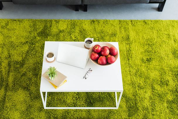 elevated view of table with laptop, coffee cups, apples in bowl, books, eyeglasses and plant at home - Fotó, kép