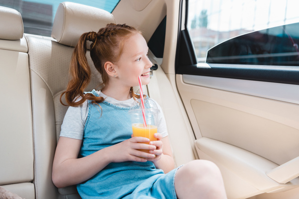 cheerful kid with juice looking out car window while sitting in car - Foto, Imagem
