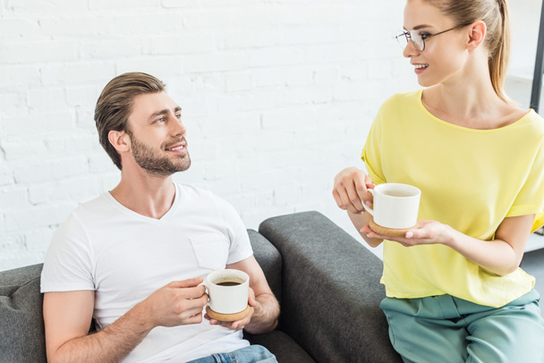 smiling young couple having conversation and drinking coffee from cups at home - Foto, Imagen