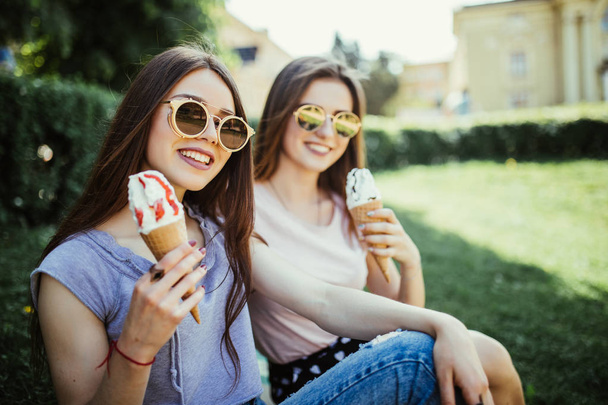 Young two women friends eat ice cream sitting on the grass in city streets - Фото, изображение