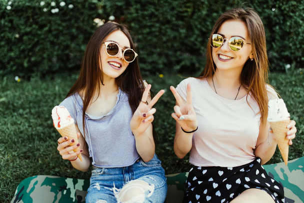 Портрет двох молодих жінок, що стоять разом, їдять морозиво, сидячи на траві на вулиці міста
. - Фото, зображення