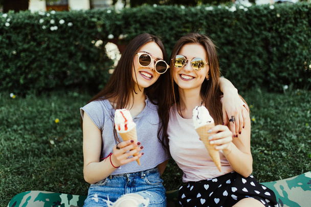 Portrait of two young women standing together eating ice cream sitting on the grass in city street. - Photo, Image