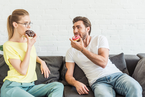Młoda para jeść pączki i siedzi na kanapie w domu  - Zdjęcie, obraz