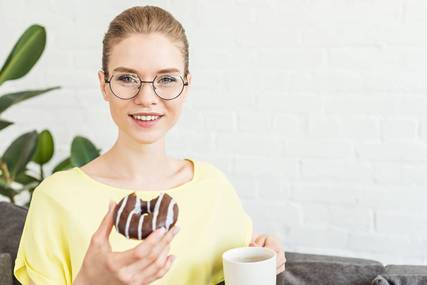 smiling young woman in eyeglasses drinking coffee and eating donut at home - Fotoğraf, Görsel