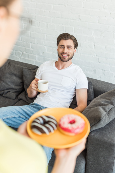 cropped shot of woman with donuts on plate and smiling boyfriend sitting on sofa with coffee cup at home - Zdjęcie, obraz