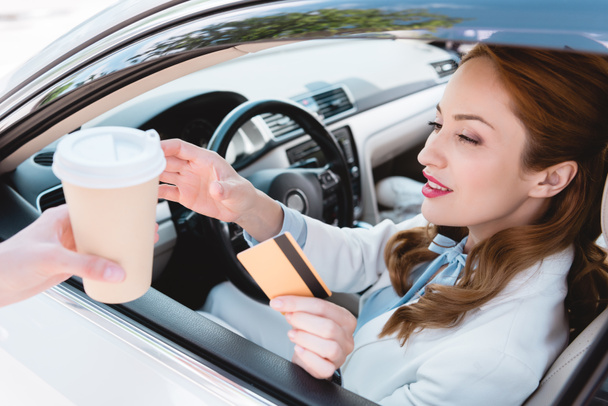 businesswoman with credit card in hand taking coffee to go while sitting in car - Photo, Image