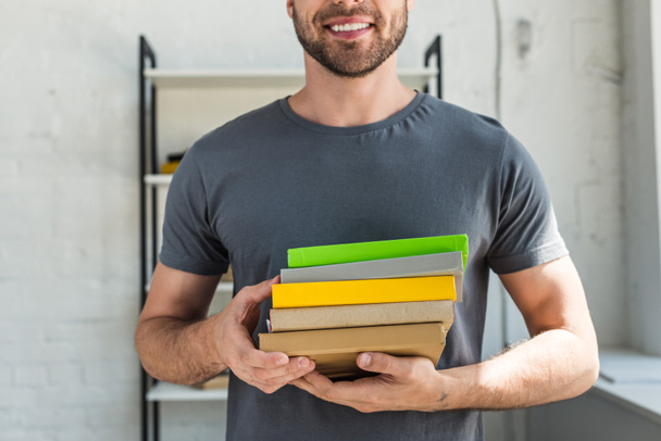 περικομμένη εικόνα της χαμογελαστός άνθρωπος στέκεται με στοίβα από βιβλία στο σπίτι  - Φωτογραφία, εικόνα