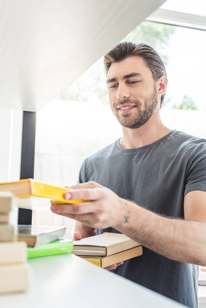 young smiling man putting books on shelves at home - Foto, Bild