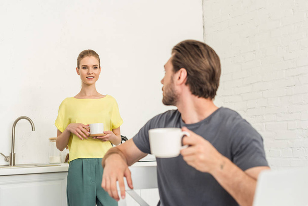 young man with coffee cup sitting on chair and talking to girlfriend at kitchen - Photo, image
