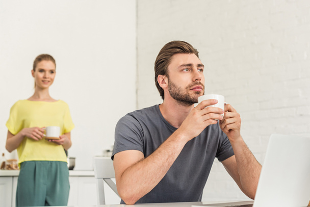 вдумчивый молодой человек сидит с чашкой кофе за столом с ноутбуком и его подругой, стоящей позади на кухне
  - Фото, изображение