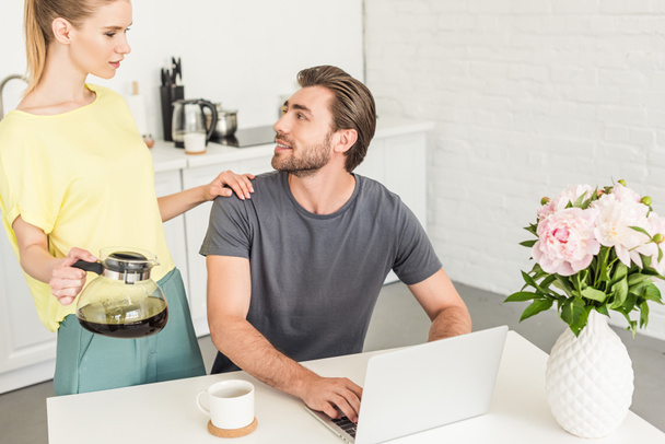 attractive young woman with coffee touching boyfriend shoulder while he sitting at table with laptop  - Foto, imagen
