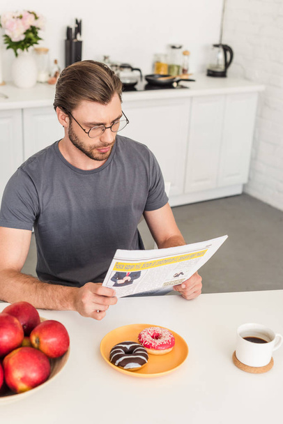 young man in eyeglasses reading newspaper at table with apples, donuts and coffee cup - 写真・画像