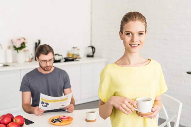 attractive smiling woman holding coffee cup and her boyfriend sitting at table and reading newspaper at kitchen  - Photo, Image