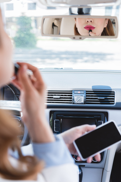partial view of businesswoman with smartphone in hand applying lipstick while driving car - Photo, Image