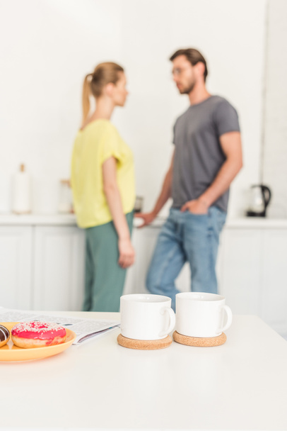 close up view of two coffee cups at table with donuts and couple standing behind at kitchen - Photo, Image