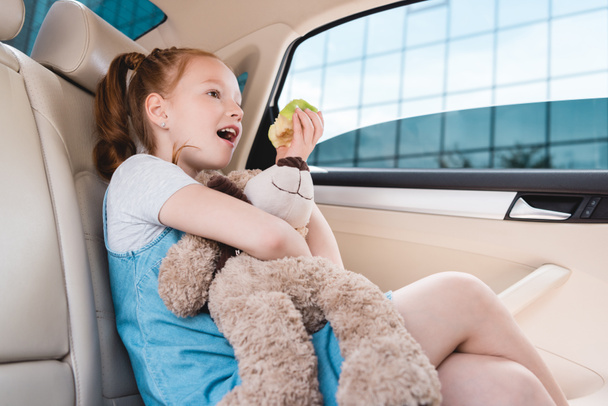 side view of emotional kid with teddy bear and fresh apple in car - Fotó, kép