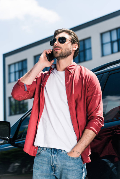 黒い車近くのスマート フォンで話しているサングラスでスタイリッシュな男性モデルの低角度のビュー  - 写真・画像