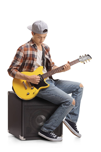 Teenager with an electric guitar sitting on an amplifier and playing isolated on white background - Foto, afbeelding