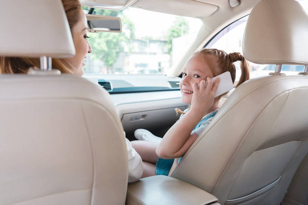 side view of smiling kid talking on smartphone with mother near by in car - Foto, afbeelding