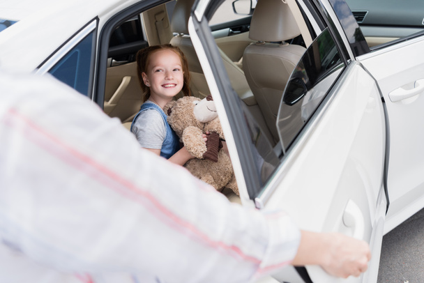 Arabanın kapısını açarak annesine bakarak bir oyuncak ayı ile gülümseyen çocuk seçici odak - Fotoğraf, Görsel