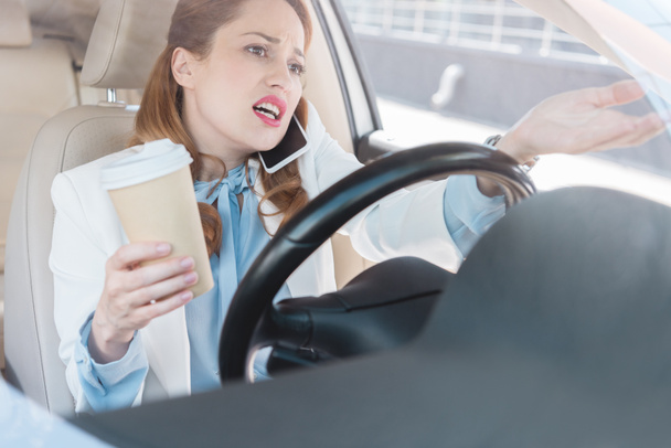 zakenvrouw met kop koffie in de hand hebben op smartphone tijdens het auto rijden - Foto, afbeelding