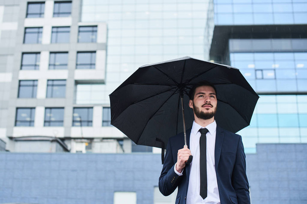 handsome bearded businessman in suit with black umbrella standing at street  - Foto, Imagen