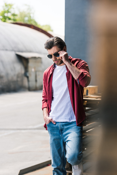 front view of young stylish man standing near wooden pallets and adjusting sunglasses  - Fotografie, Obrázek