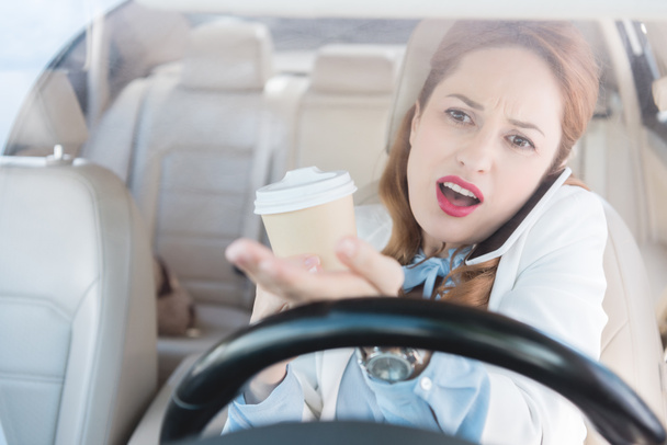 businesswoman with cup of coffee in hand talking on smartphone while driving car - Fotoğraf, Görsel